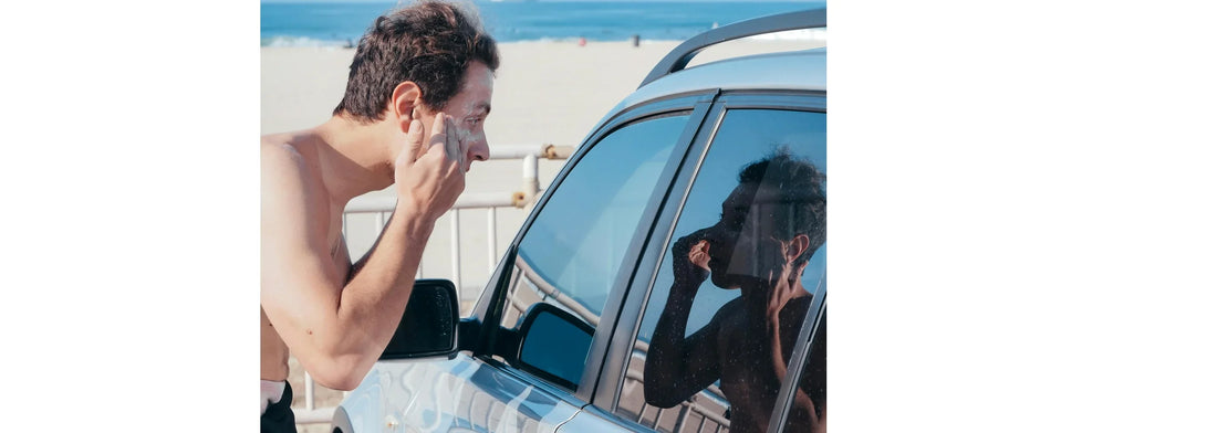 Men's Skin Care Tips That Actually Work For Summer photo of man at the beach putting on sun screen and looking into a car window so he can see what he is doing.